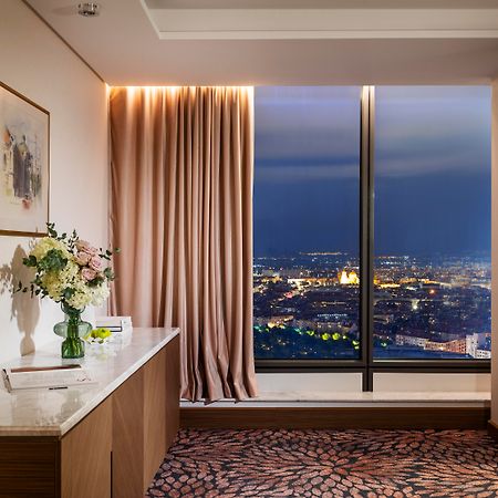 Grand Hotel Millennium Софія Екстер'єр фото The photo depicts a modern hotel room or a lounge area with a stunning view of a cityscape at night. On the left, there's a wooden console table with a vase of flowers and a bottle of water. A piece of art is framed on the wall above the table. The r