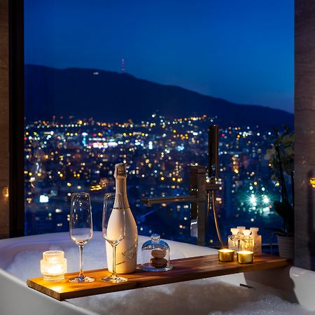 Grand Hotel Millennium Софія Екстер'єр фото The photo features a luxurious bathtub placed by a large window, showcasing a scenic city view at night. There is a wooden tray resting on the edge of the tub, displaying a bottle of champagne, two glass flutes, and several lit candles, creating an a