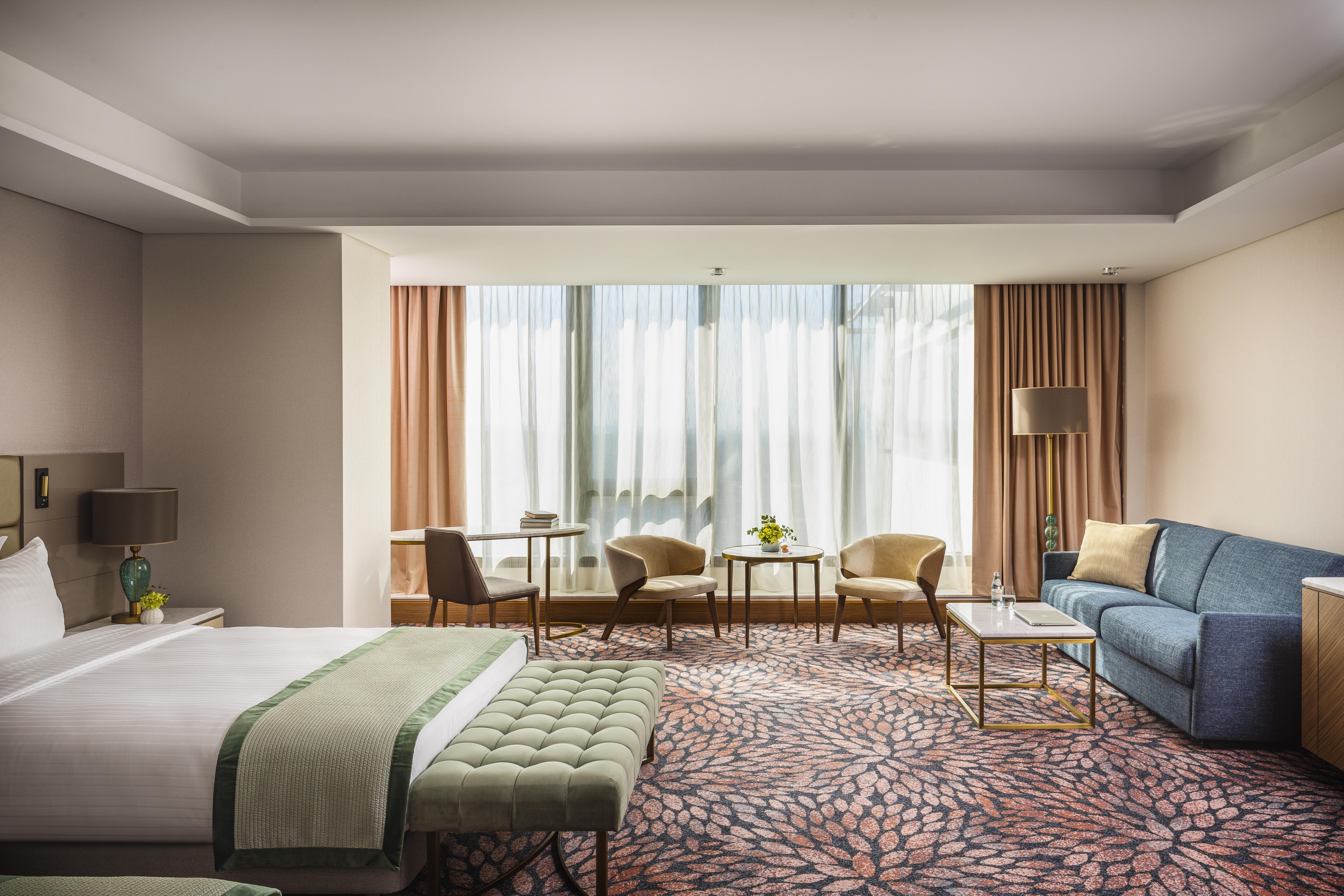 Grand Hotel Millennium Софія Екстер'єр фото The photo shows a modern hotel room featuring a spacious layout. On one side, there is a large bed with a green throw blanket and a tufted bench at its foot. The room is well-lit by large windows dressed with light curtains, which allow natural light