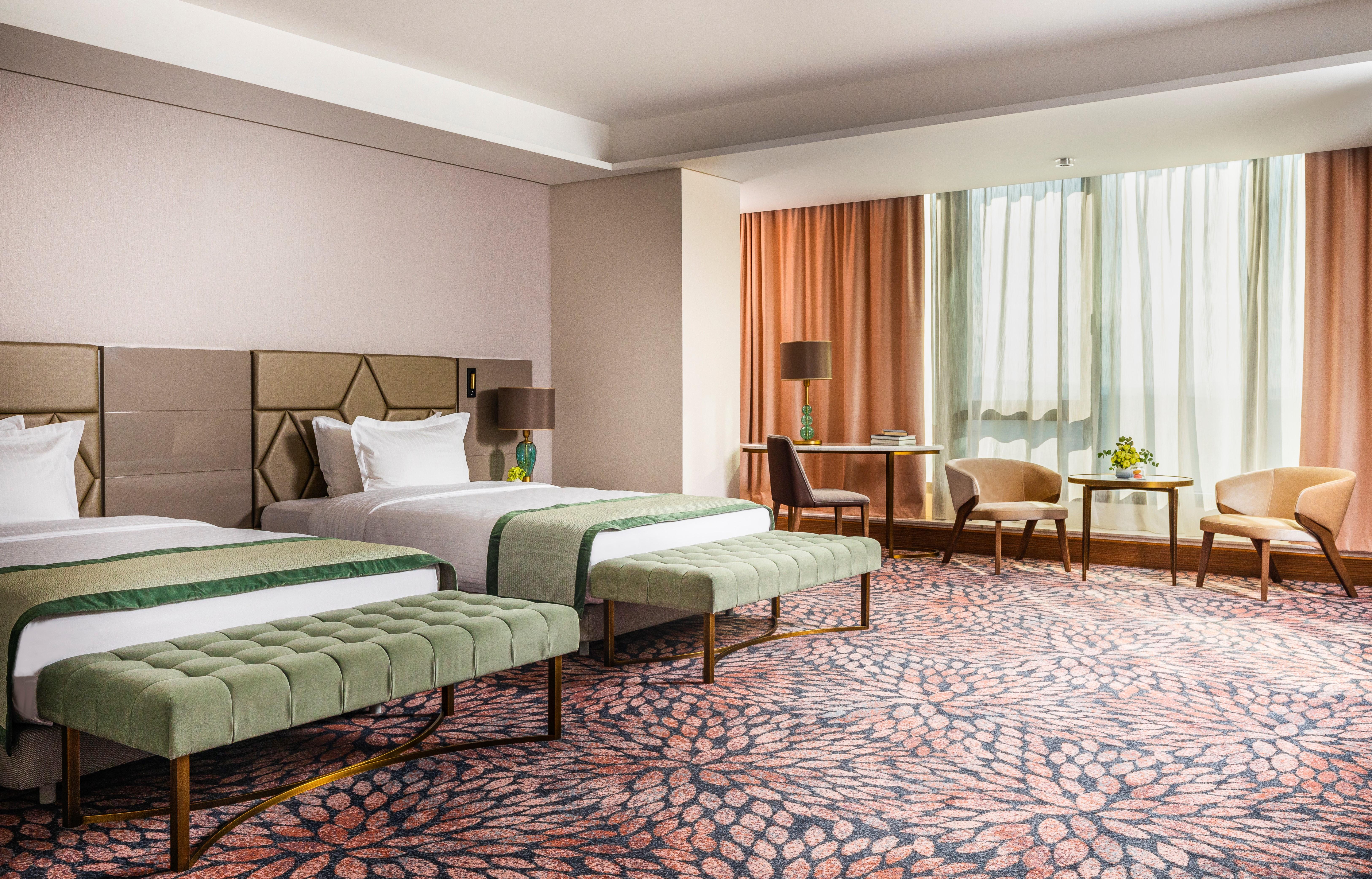 Grand Hotel Millennium Софія Екстер'єр фото The image shows a well-decorated hotel room featuring two beds with green striped bedspreads. There is a plush green bench at the foot of each bed. The room has large windows with sheer curtains allowing natural light to enter. There is a cozy seatin