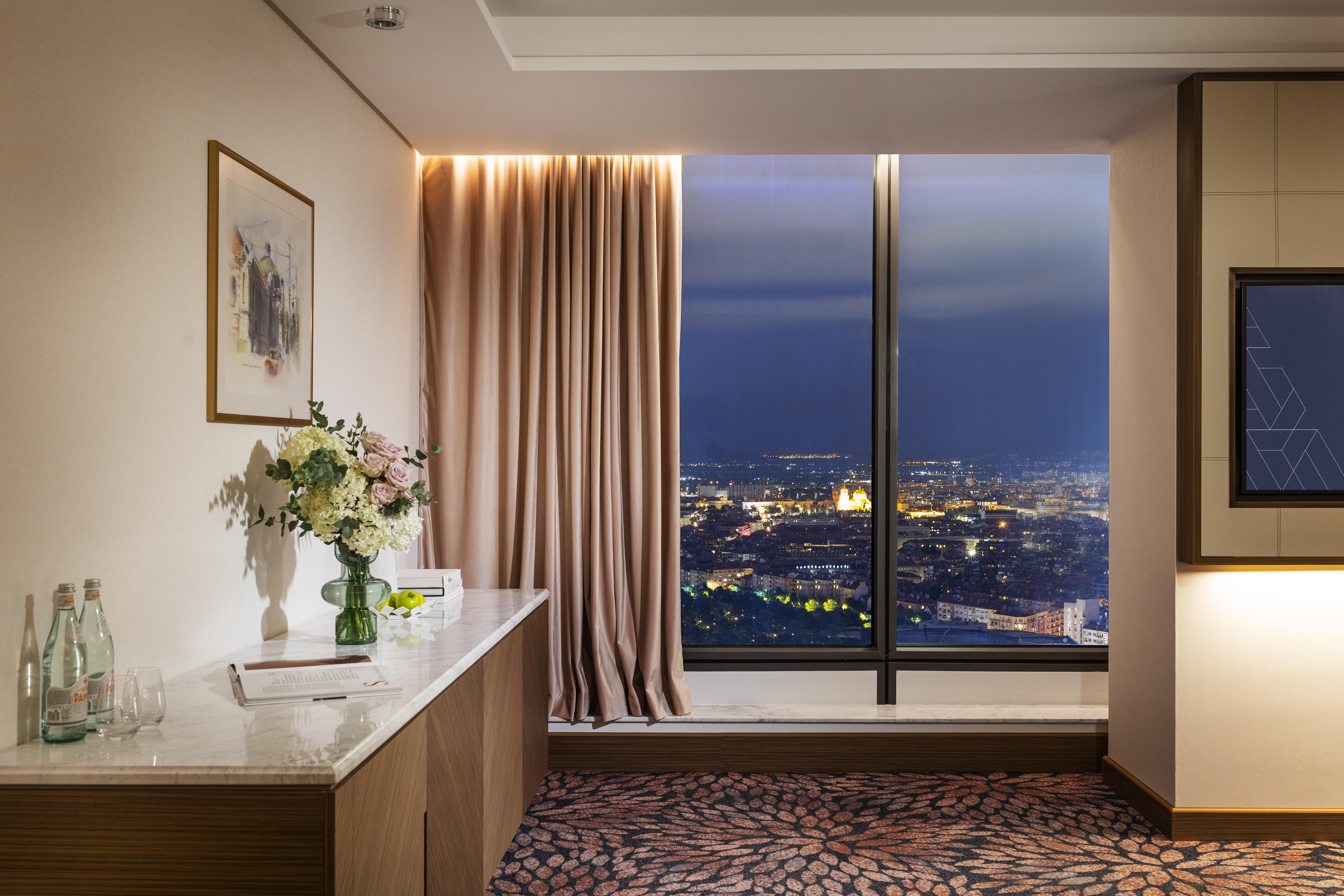 Grand Hotel Millennium Софія Екстер'єр фото The photo depicts a modern hotel room or a lounge area with a stunning view of a cityscape at night. On the left, there's a wooden console table with a vase of flowers and a bottle of water. A piece of art is framed on the wall above the table. The r