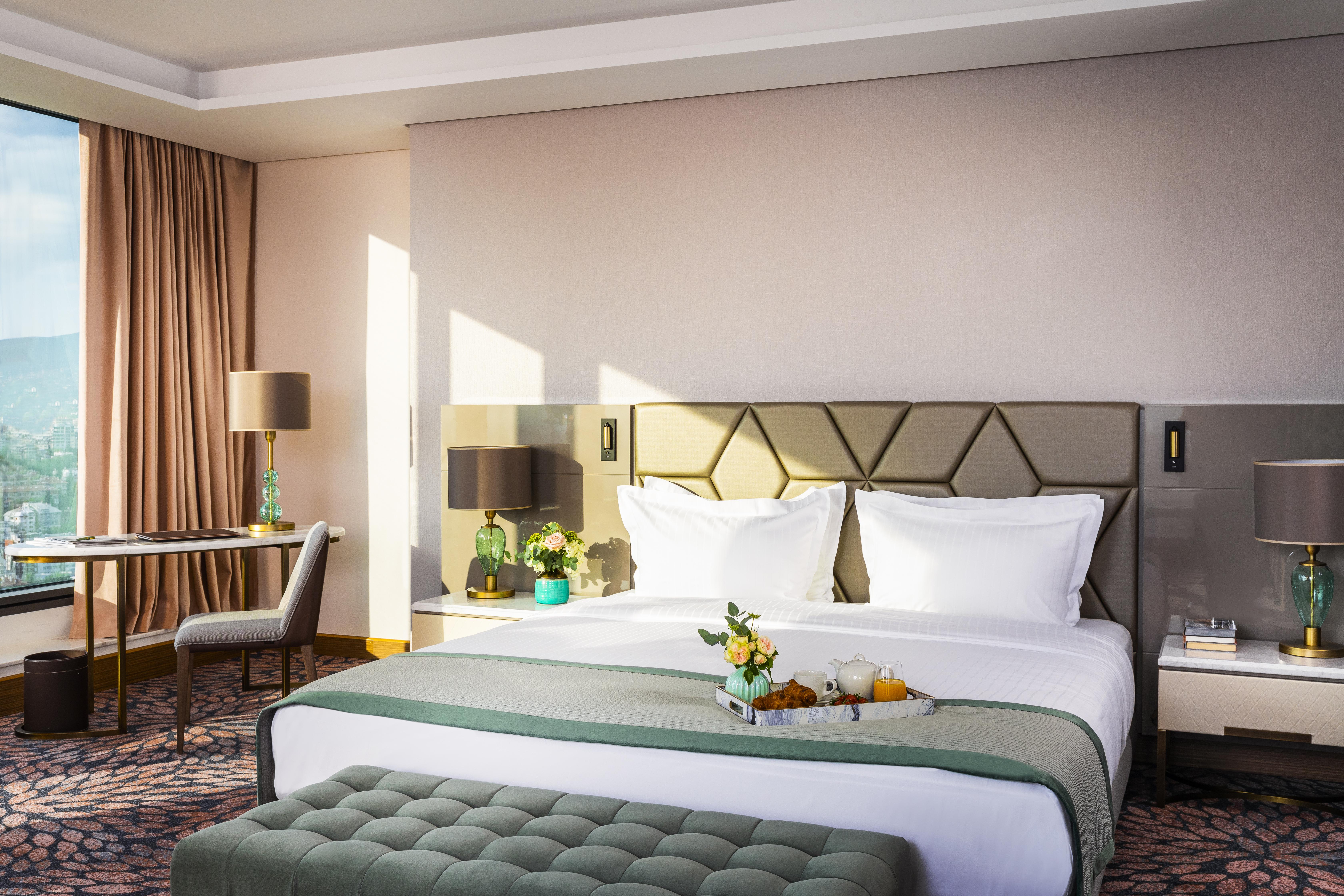 Grand Hotel Millennium Софія Екстер'єр фото The photo depicts a stylish hotel room featuring a large bed with white linens and a green decorative throw. Behind the bed is a geometric-patterned headboard. To the side, there are modern bedside lamps and a small table with a vase of flowers. A se