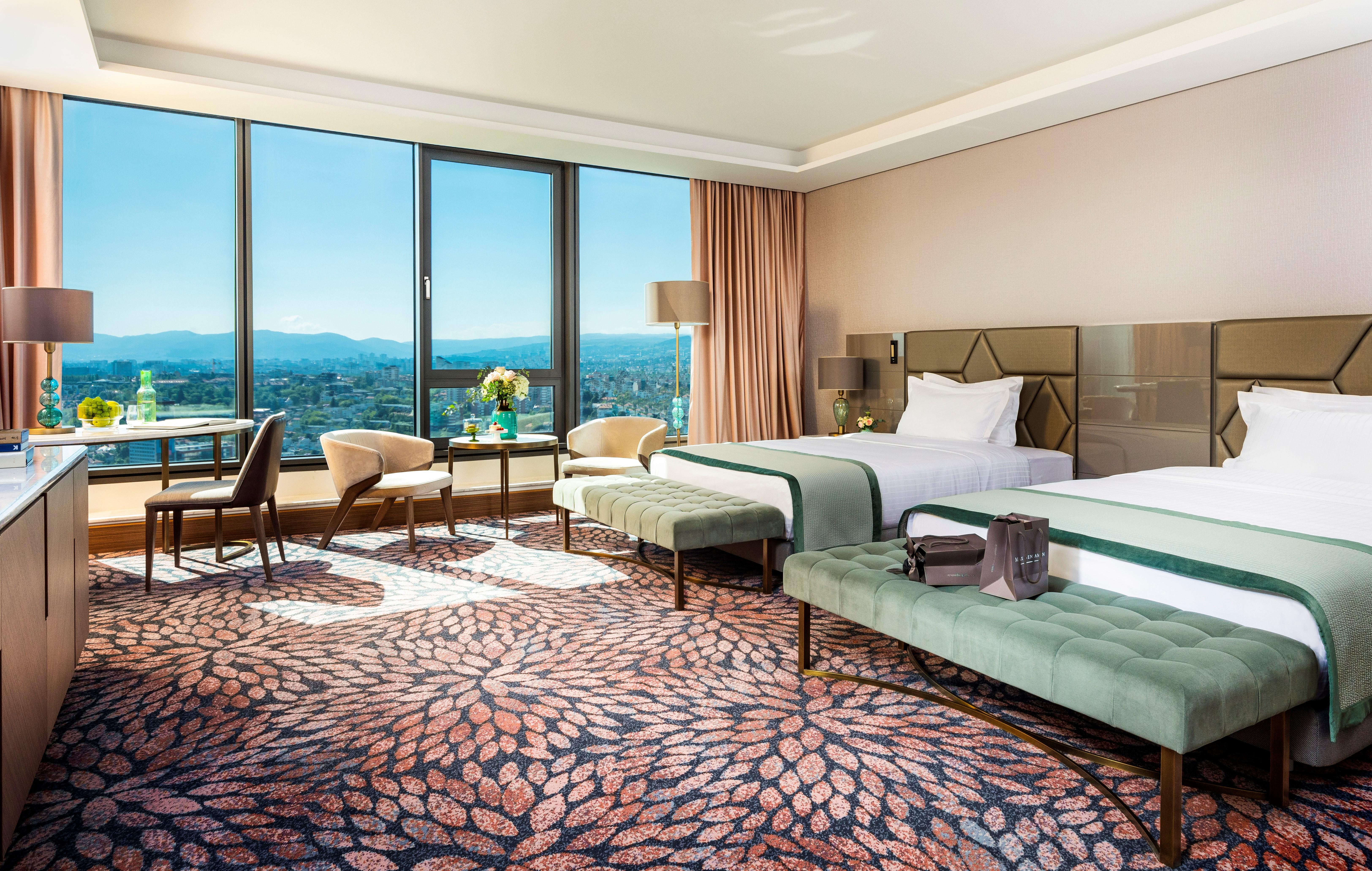 Grand Hotel Millennium Софія Екстер'єр фото The photo depicts a luxurious hotel room featuring two beds with elegant bedding. The room has large windows that offer a panoramic view of the city and mountains in the background. There is a decorative area rug with a floral pattern covering the fl