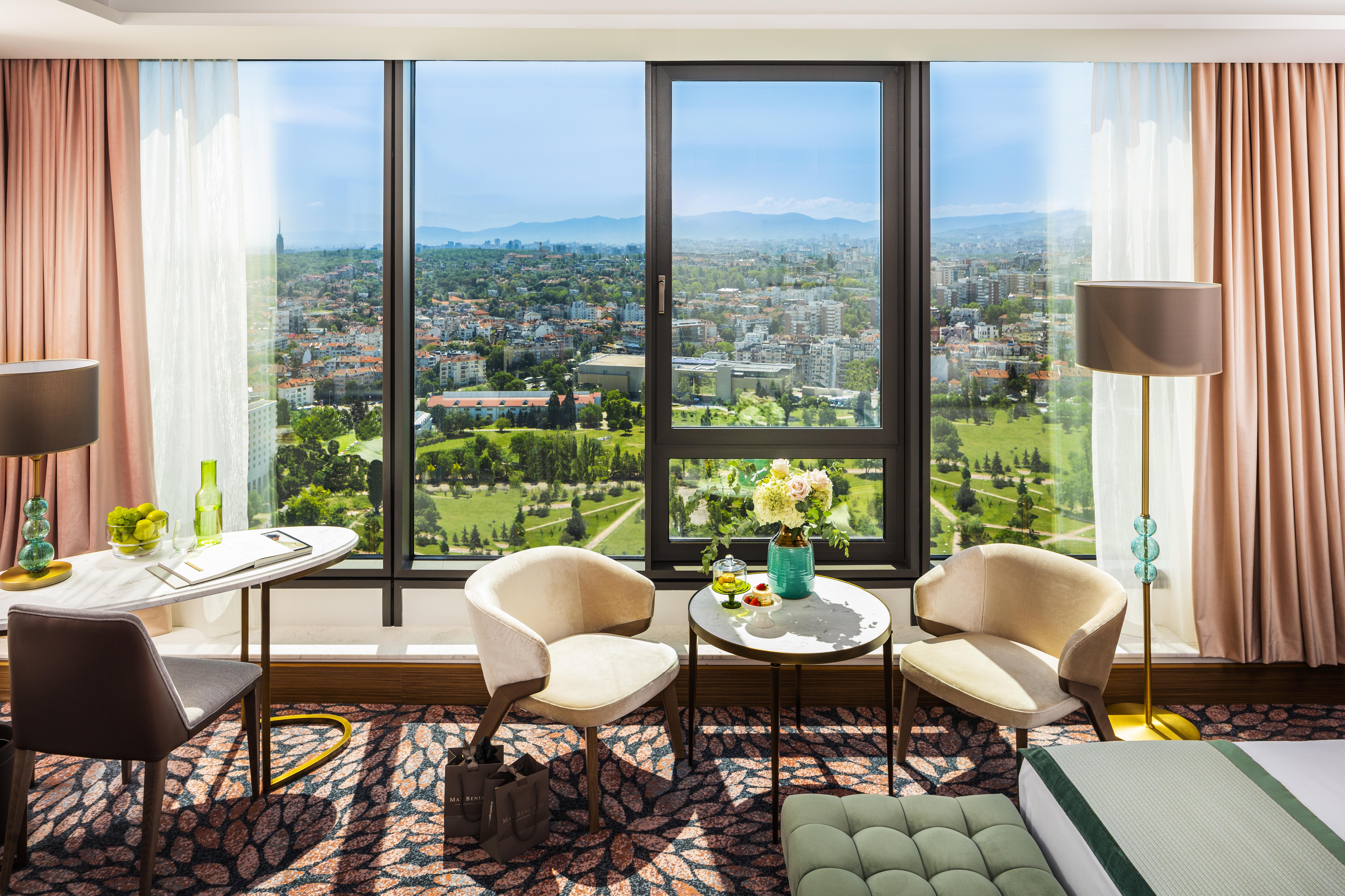 Grand Hotel Millennium Софія Екстер'єр фото The photo shows a stylish hotel room with large windows providing a beautiful view of a cityscape and landscape beyond. There are two modern chairs and a small round table set near the windows, accompanied by a vase of fresh flowers. The room feature