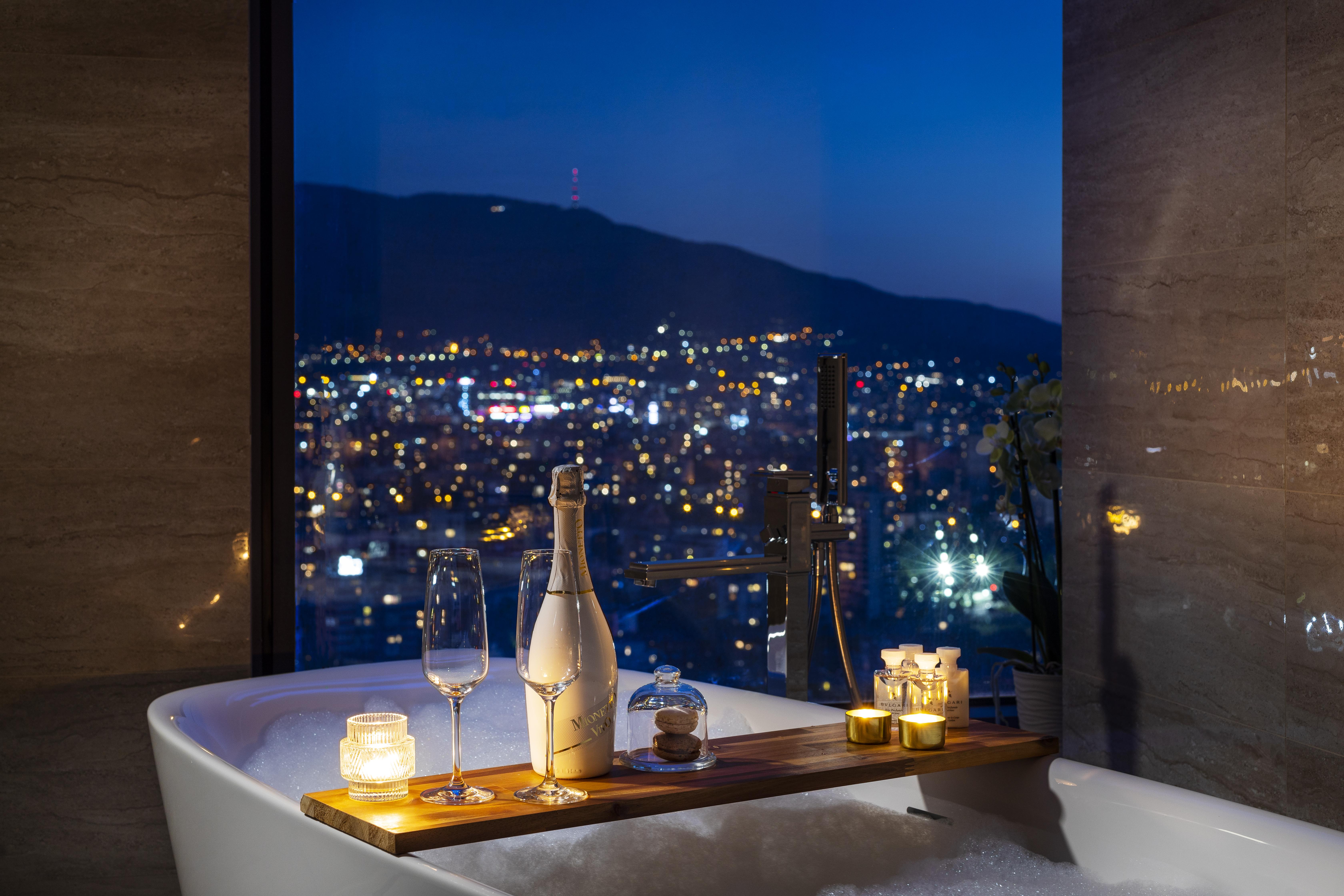 Grand Hotel Millennium Софія Екстер'єр фото The photo features a luxurious bathtub placed by a large window, showcasing a scenic city view at night. There is a wooden tray resting on the edge of the tub, displaying a bottle of champagne, two glass flutes, and several lit candles, creating an a