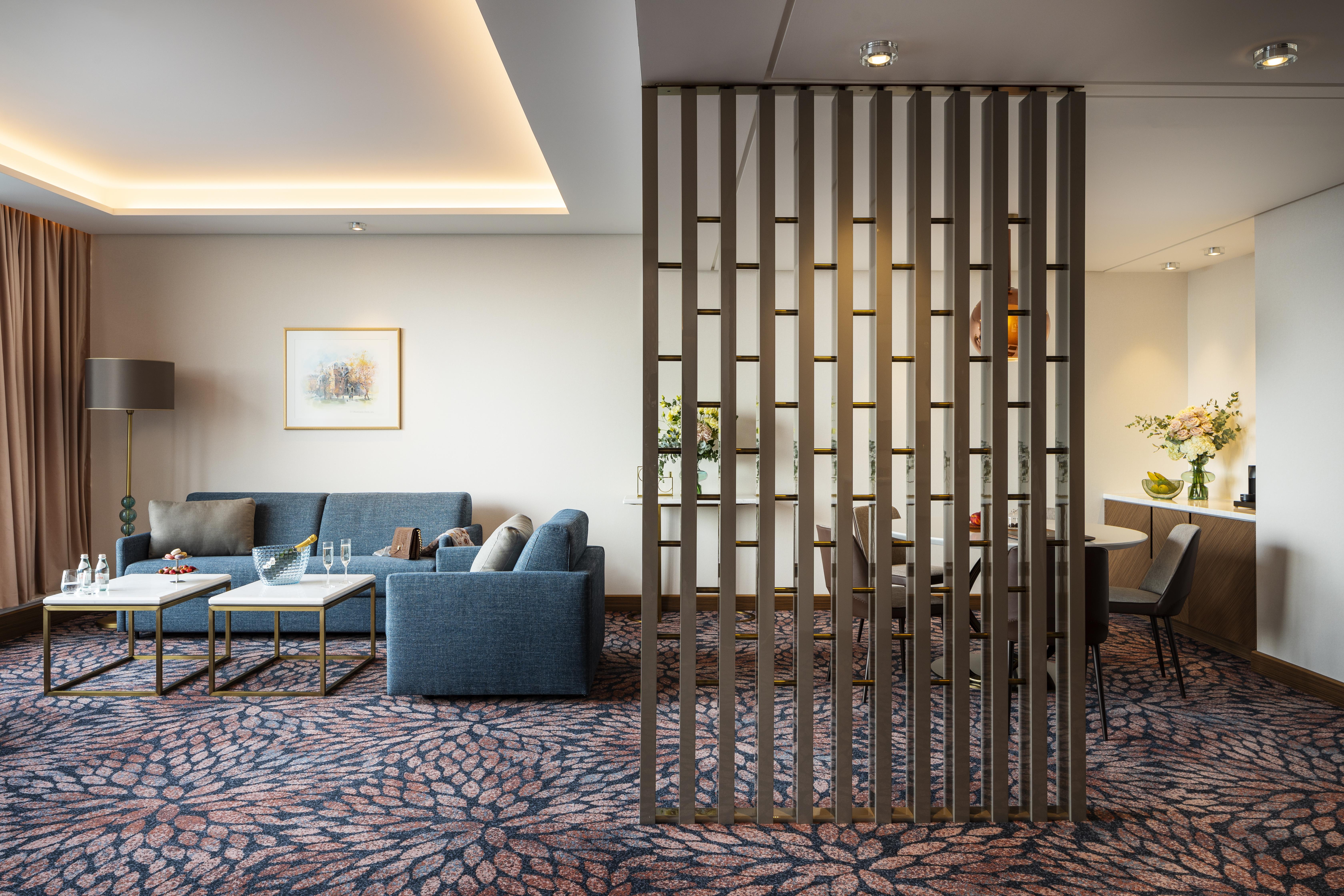 Grand Hotel Millennium Софія Екстер'єр фото The photo shows a modern living space with a comfortable seating area featuring a blue couch and two coffee tables. There is a decorative partition made of vertical wooden slats, which separates this sitting area from a dining space. The dining area 