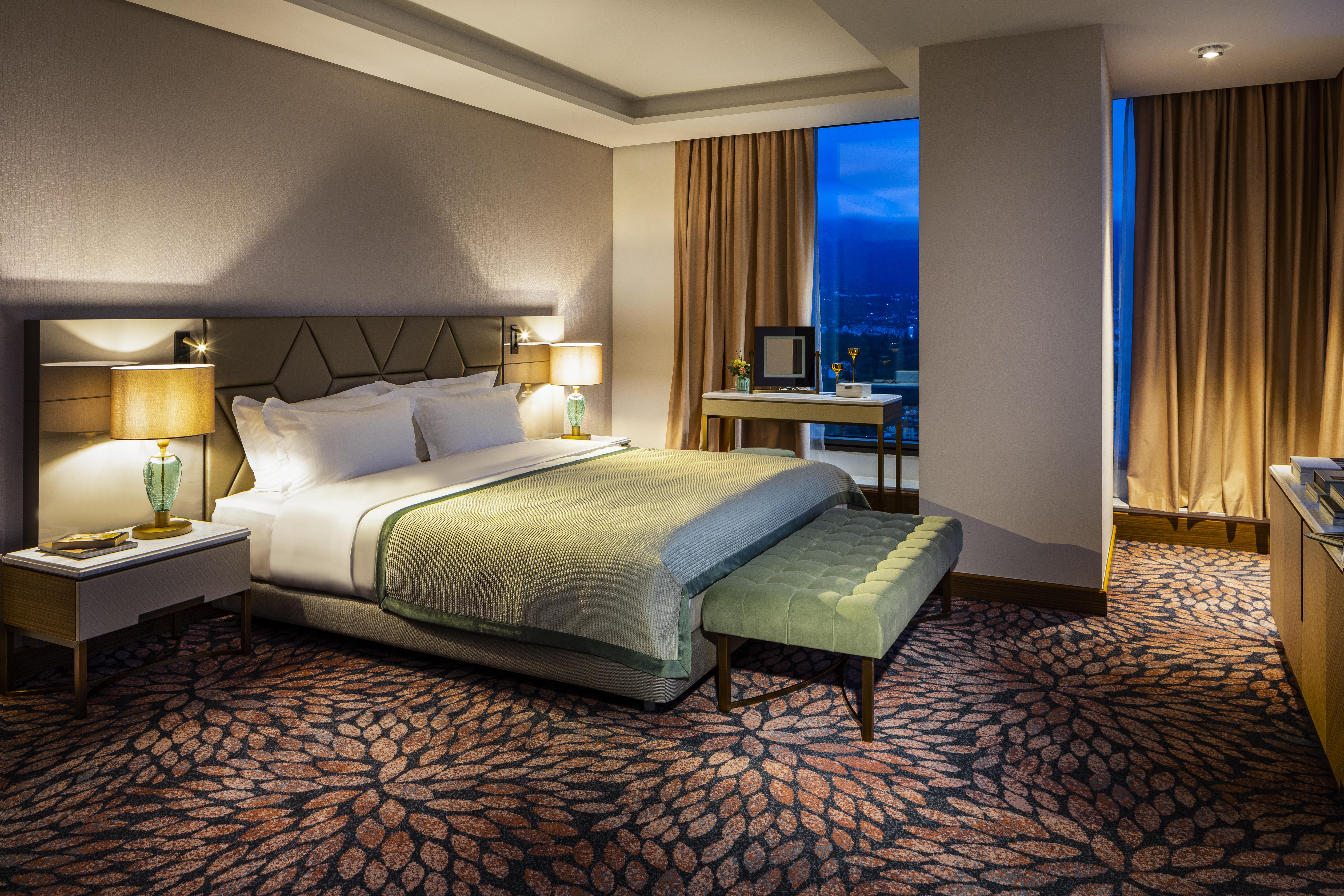 Grand Hotel Millennium Софія Екстер'єр фото The photo shows a modern hotel room featuring a spacious design. It has a large bed with a light green throw blanket positioned in the center. On either side of the bed, there are stylish bedside tables with lamps. 

The floor has a patterned carpet,