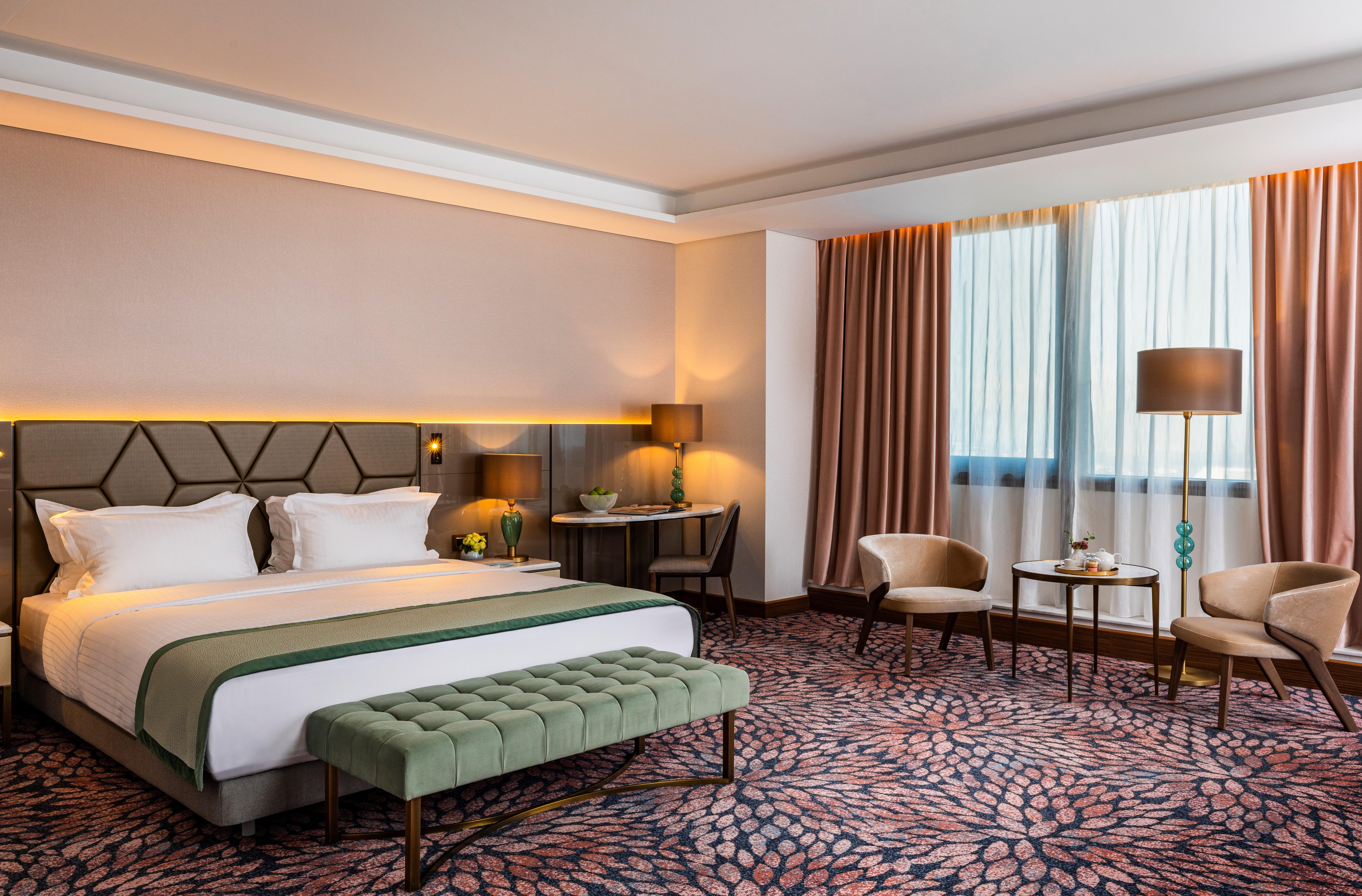 Grand Hotel Millennium Софія Екстер'єр фото The photo shows a modern hotel room. It features a large bed with a headboard, topped with two white pillows and a green throw blanket. In front of the bed, there is a stylish green bench. On either side of the bed, there are bedside tables with lamp
