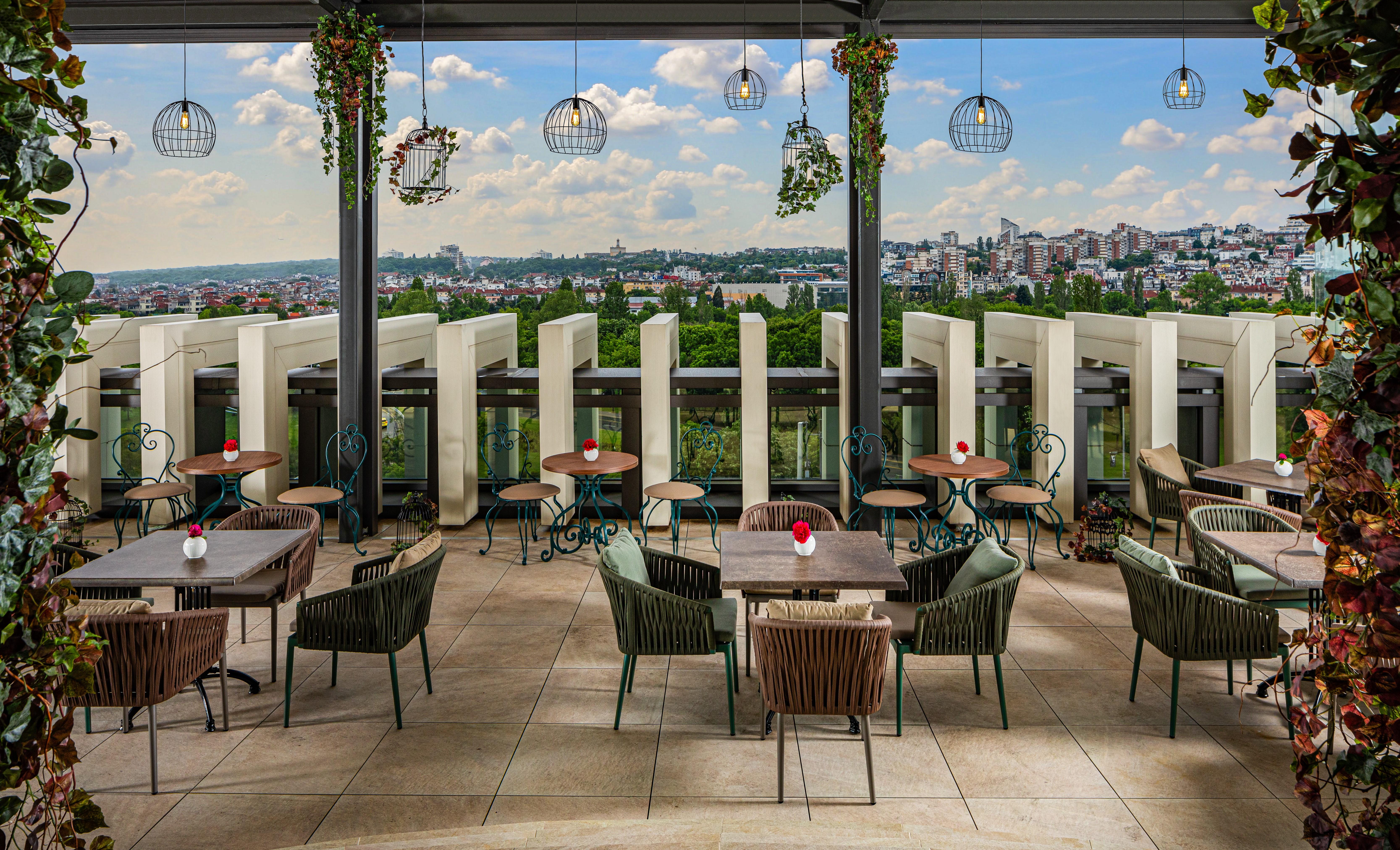 Grand Hotel Millennium Софія Екстер'єр фото The photo shows a stylish outdoor restaurant or café setting. There are several tables arranged on a tiled patio, each with chairs, some made of woven materials. Centerpieces with small red flowers adorn each table. The space is partially shaded by o