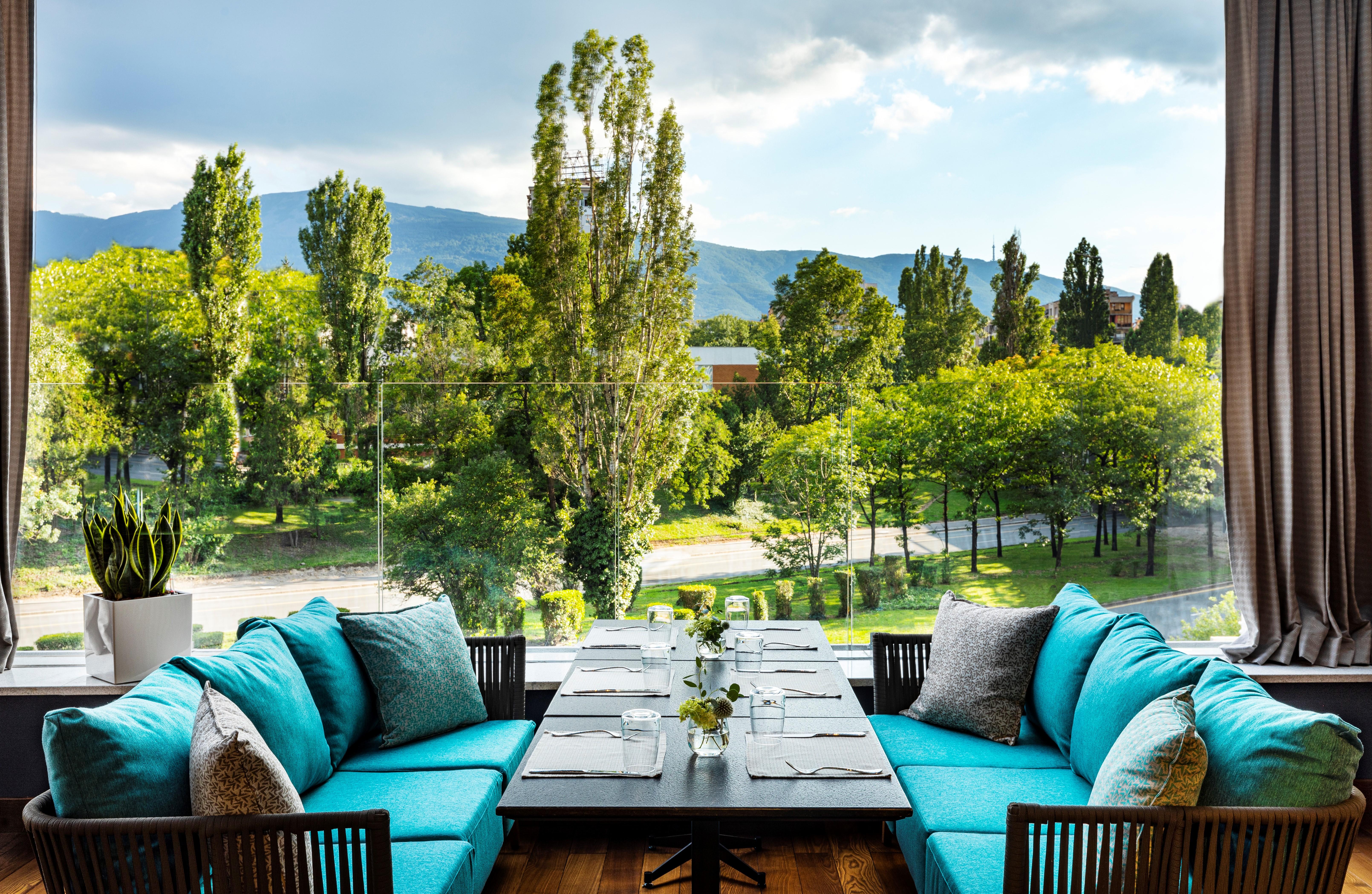 Grand Hotel Millennium Софія Екстер'єр фото The photo shows a bright and inviting dining area with large windows that overlook a lush green landscape. There are several trees in the background, along with a view of distant mountains under a partly cloudy sky. The seating consists of stylish tu