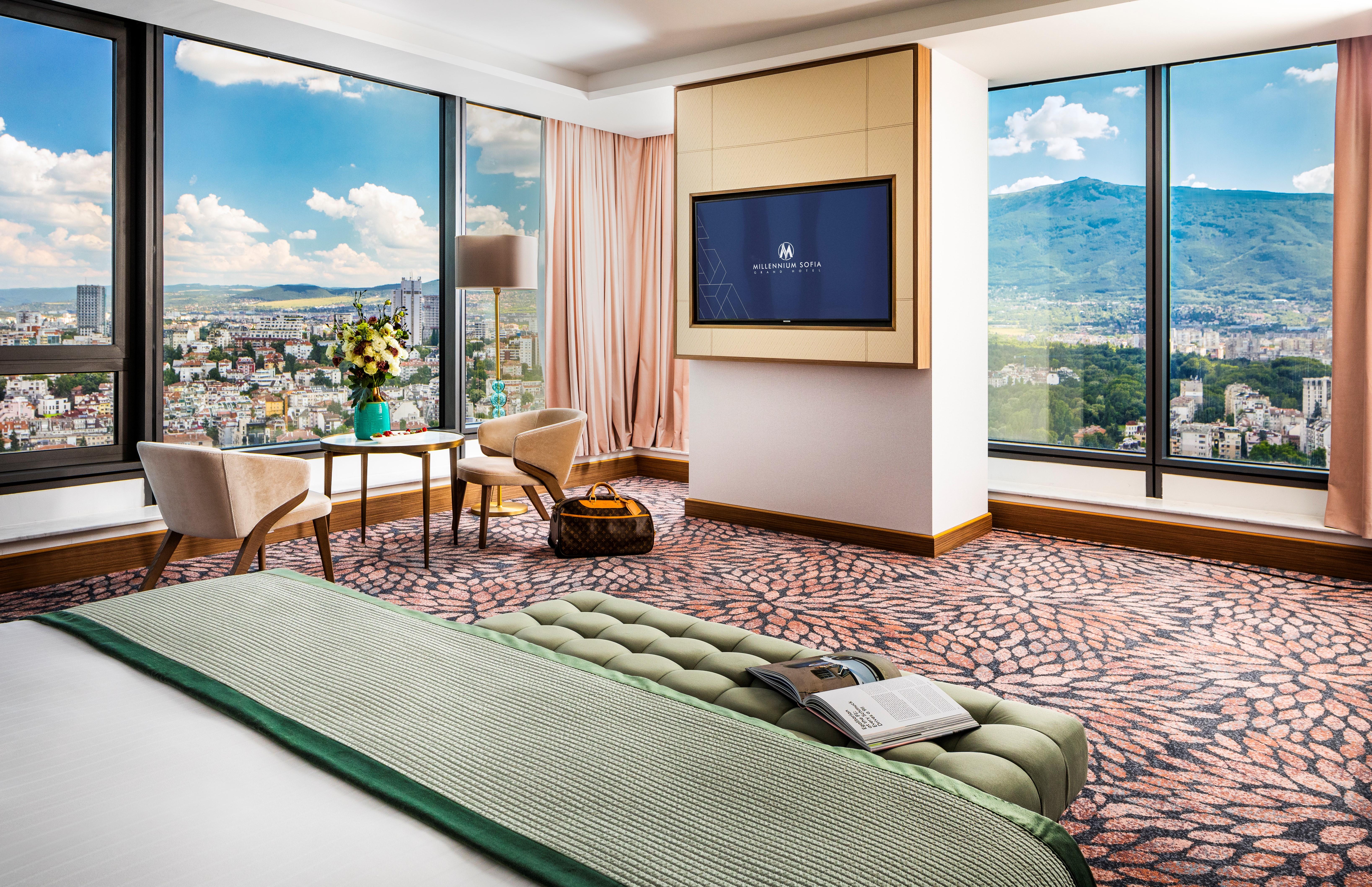 Grand Hotel Millennium Софія Екстер'єр фото The photo showcases a stylish hotel room with modern decor. It features large windows providing a panoramic view of a cityscape and mountains in the background. Inside, there is a comfortable bed with a green bedspread, a seating area with two armcha