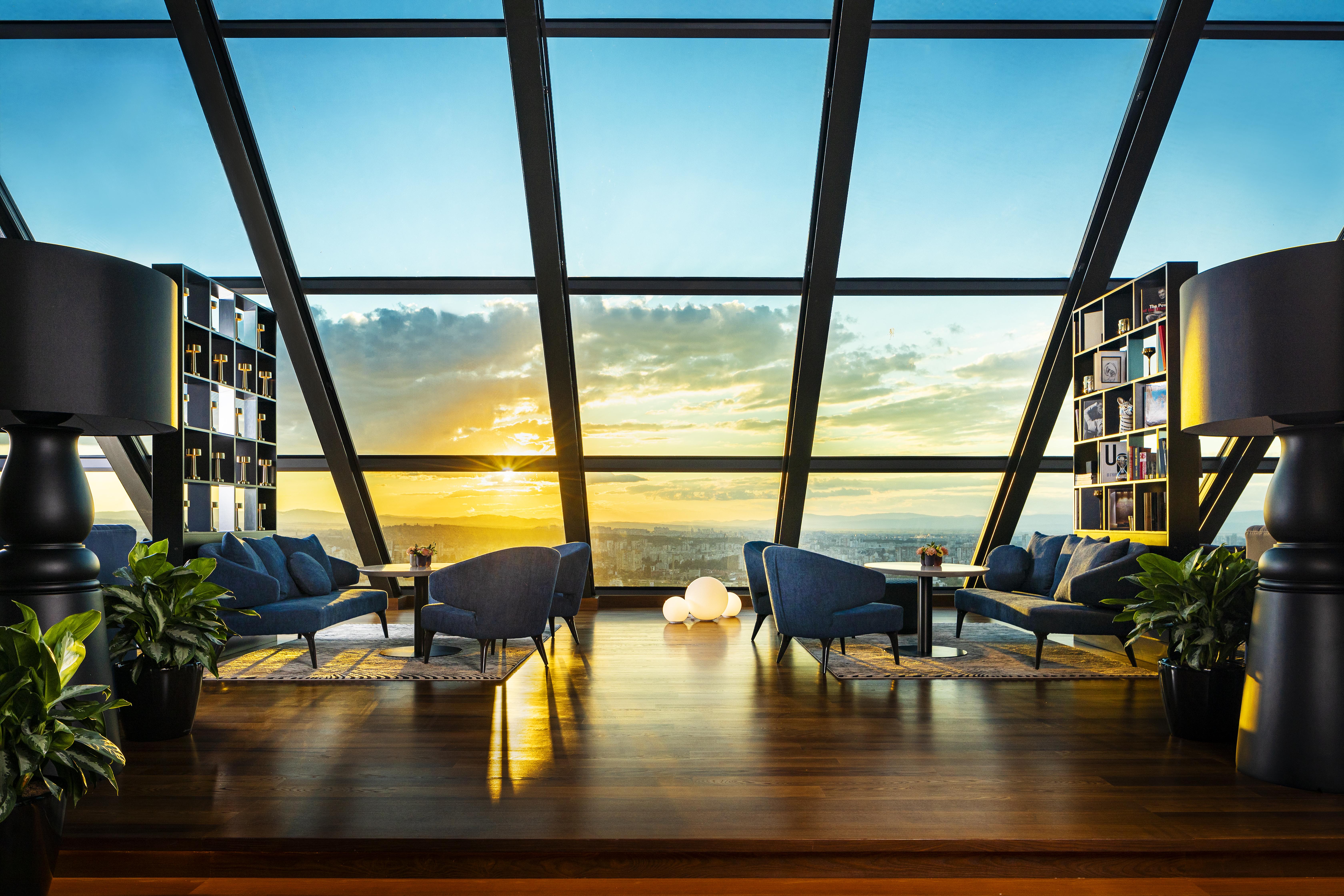 Grand Hotel Millennium Софія Екстер'єр фото The photo depicts a modern, stylish interior of a high-rise space with a panoramic view through large glass windows. The scene features a cozy seating area with several blue armchairs arranged around a low table, creating a comfortable atmosphere. Th