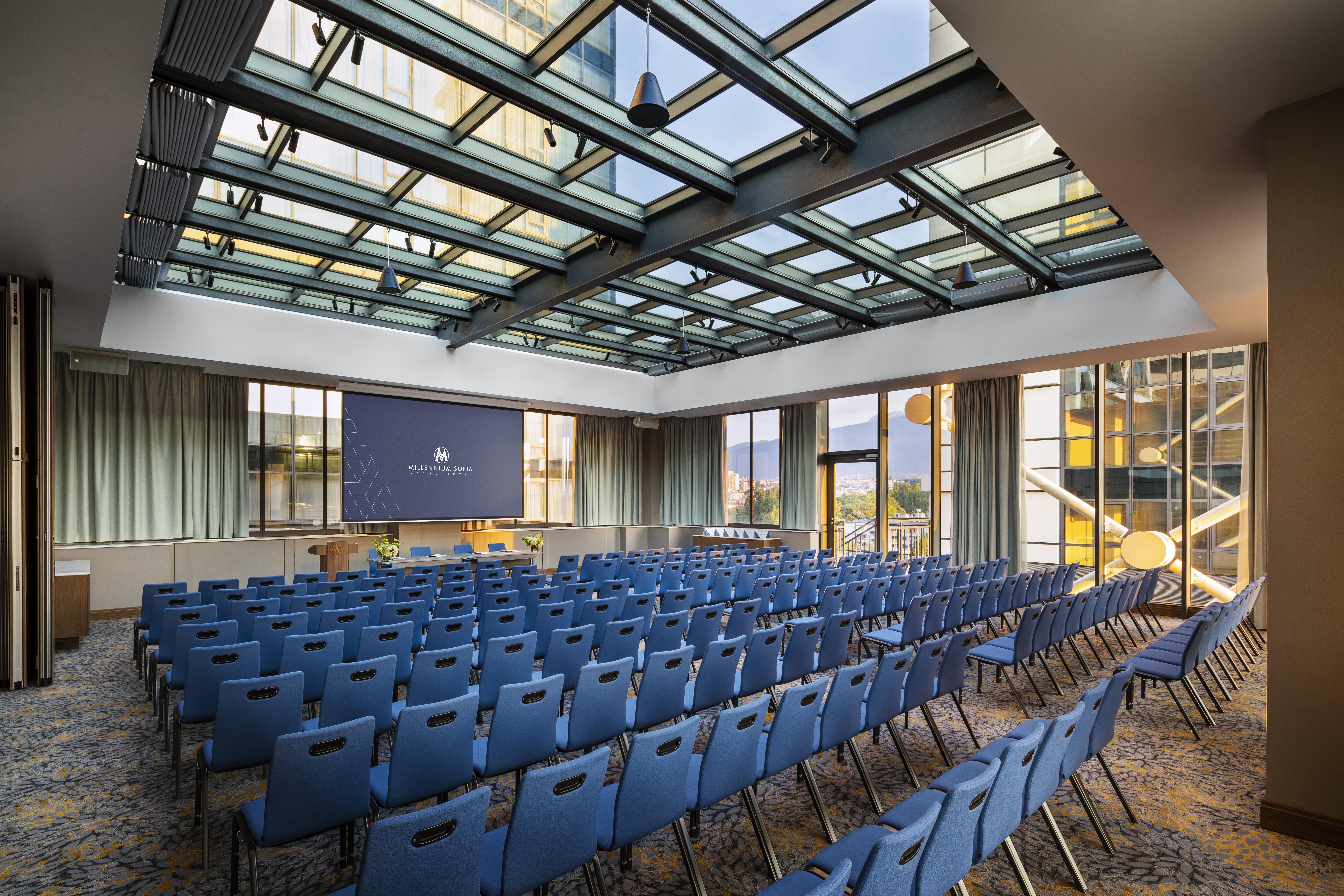 Grand Hotel Millennium Софія Екстер'єр фото The photo shows a large conference room or meeting space. It features rows of blue chairs arranged in a theater style, facing a large screen at the front of the room. The room is well-lit, with a glass ceiling allowing natural light to filter in. The