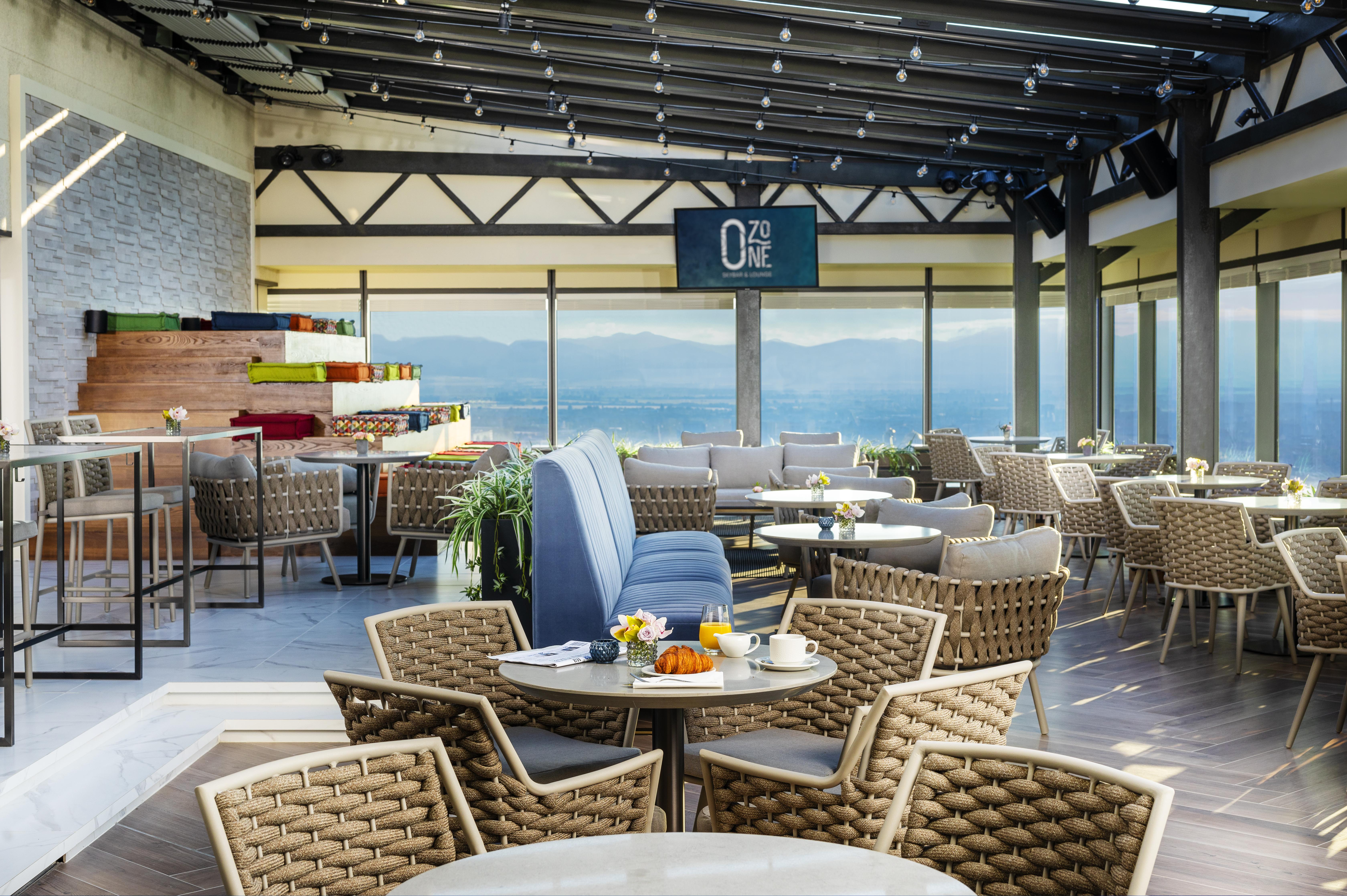 Grand Hotel Millennium Софія Екстер'єр фото The photo shows a modern restaurant or café with a spacious interior. It features a variety of seating arrangements, including round tables surrounded by wicker chairs and lounges. Large windows allow for a panoramic view, which likely includes sceni