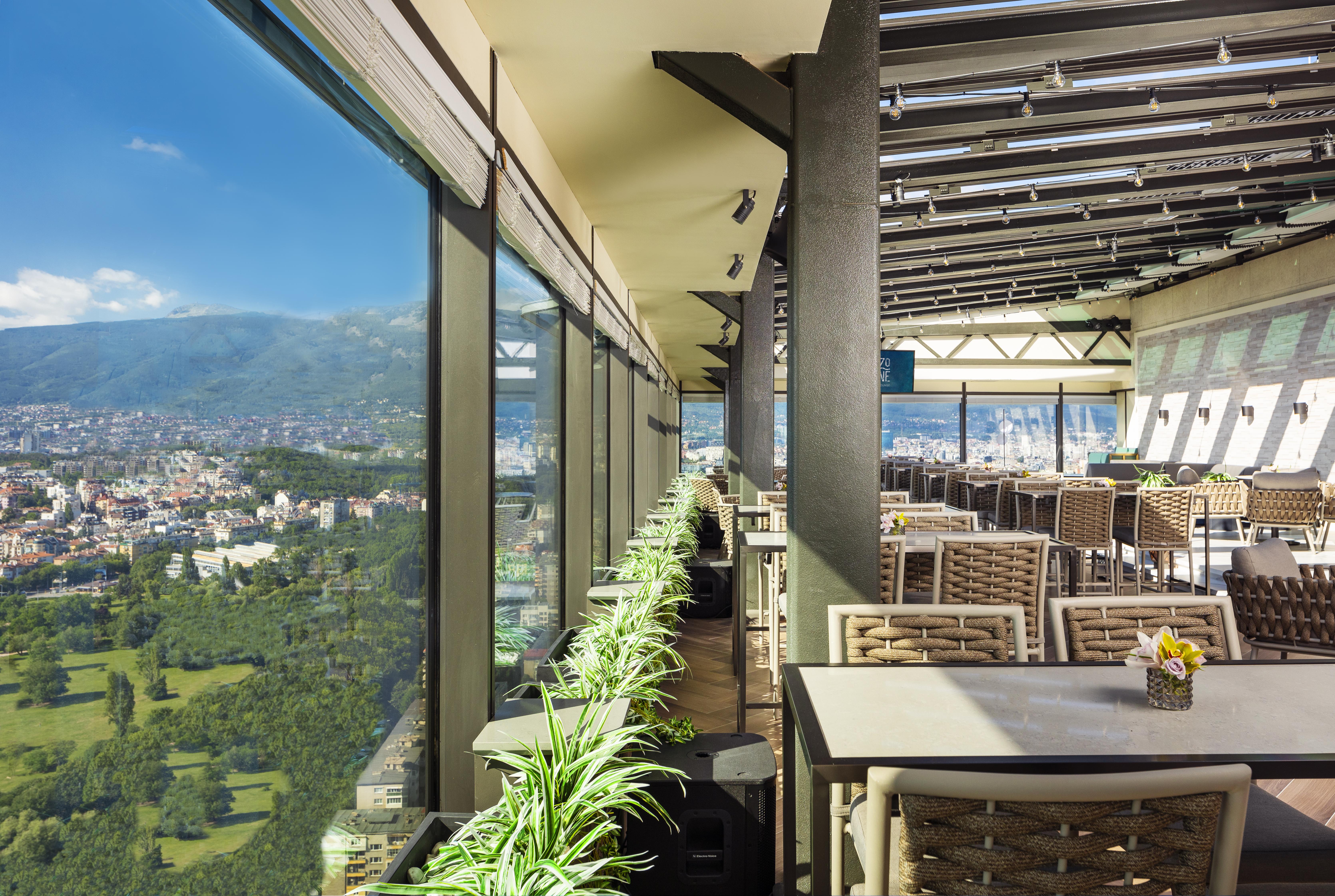 Grand Hotel Millennium Софія Екстер'єр фото The photo shows a modern, spacious restaurant with large glass windows offering a panoramic view of a city and surrounding landscape. The interior features contemporary furniture, including stylish tables and chairs. There are potted plants along the